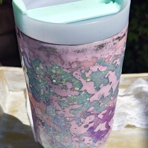 Puede incluir: Un vaso cil&iacute;ndrico con un patr&oacute;n jaspeado en tonos rosa pastel, azul claro y morado, con purpurina iridiscente incrustada en toda la superficie. El vaso tiene una tapa blanca transl&uacute;cida y una pesta&ntilde;a deslizante de color verde menta.