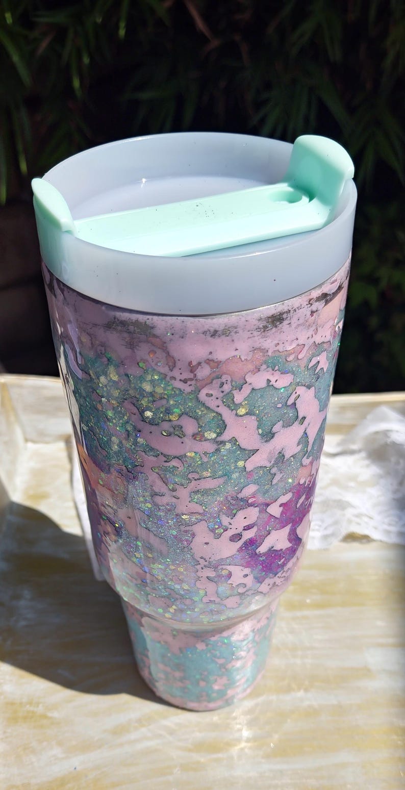 Puede incluir: Un vaso cil&iacute;ndrico con un patr&oacute;n jaspeado en tonos rosa pastel, azul claro y morado, con purpurina iridiscente incrustada en toda la superficie. El vaso tiene una tapa blanca transl&uacute;cida y una pesta&ntilde;a deslizante de color verde menta.