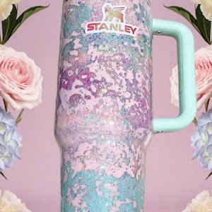 Puede incluir: Un vaso cil&iacute;ndrico alto con asa y tapa de color verde menta claro. El cuerpo presenta un acabado de purpurina texturizado y multicolor en tonos rosa, morado y azul. El logotipo de Stanley est&aacute; impreso en blanco y rojo cerca del borde.
