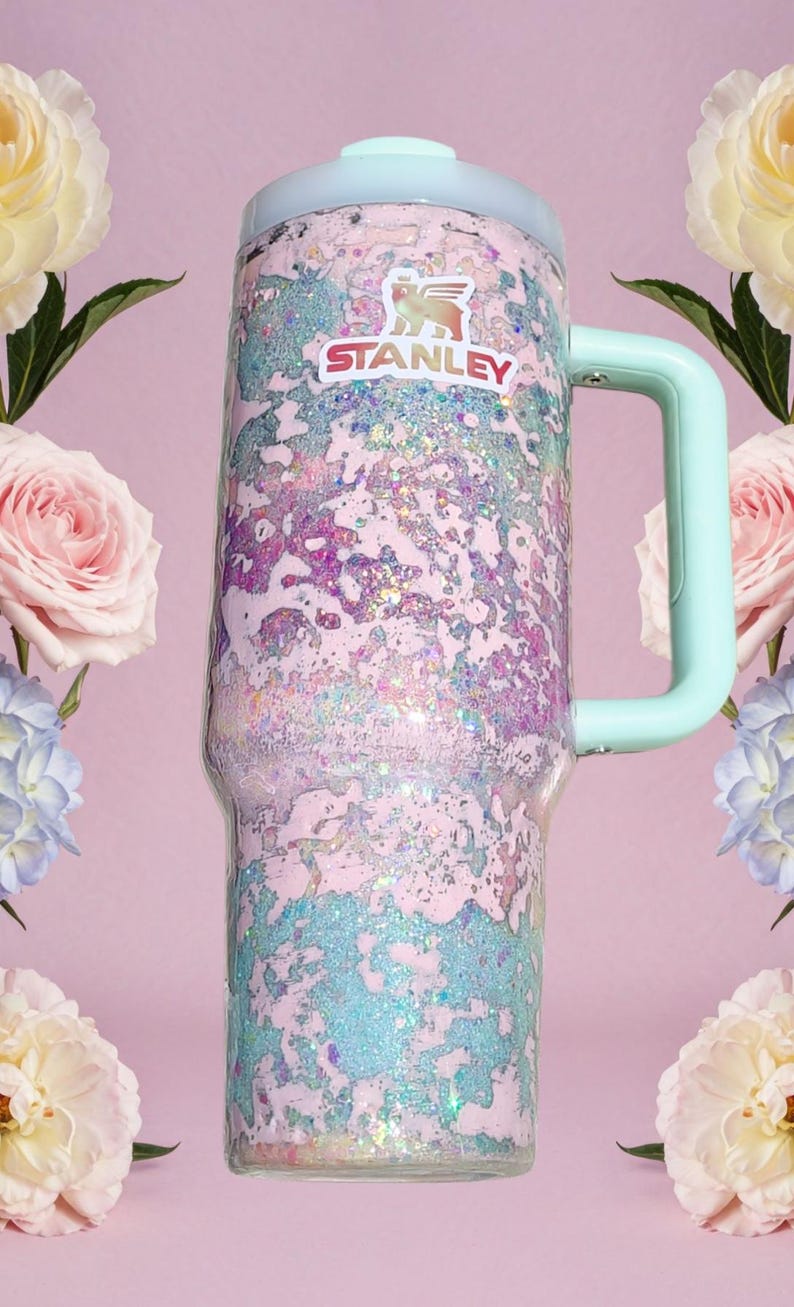 Puede incluir: Un vaso cil&iacute;ndrico alto con asa y tapa de color verde menta claro. El cuerpo presenta un acabado de purpurina texturizado y multicolor en tonos rosa, morado y azul. El logotipo de Stanley est&aacute; impreso en blanco y rojo cerca del borde.