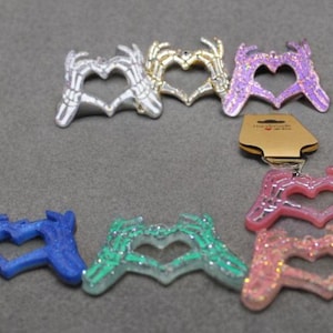 Puede incluir: Una colección de colgantes en forma de corazón en plata, oro, morado, rosa, azul y verde. Los colgantes tienen un diseño de mano de esqueleto y purpurina. Algunos tienen llaveros.