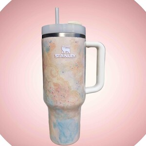 Puede incluir: Un vaso de la marca Stanley con asa y pajita. El vaso presenta un dise&ntilde;o colorido con tonos de azul, melocot&oacute;n y crema. El logotipo de Stanley es visible en la parte delantera del vaso.