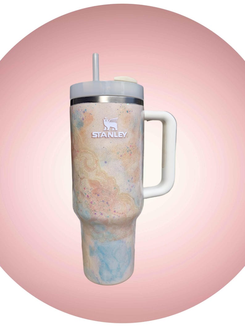 Puede incluir: Un vaso de la marca Stanley con asa y pajita. El vaso presenta un dise&ntilde;o colorido con tonos de azul, melocot&oacute;n y crema. El logotipo de Stanley es visible en la parte delantera del vaso.