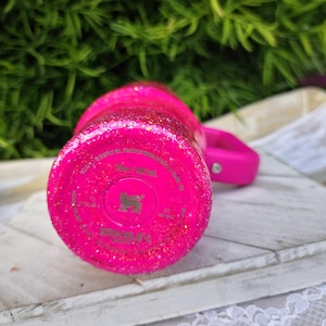 Pode incluir: Um copo rosa brilhante coberto de glitter com uma al&ccedil;a a condizer. A parte inferior do copo tem o nome e o log&oacute;tipo da marca. O copo est&aacute; sobre uma superf&iacute;cie de madeira branca com uma borda de renda branca.