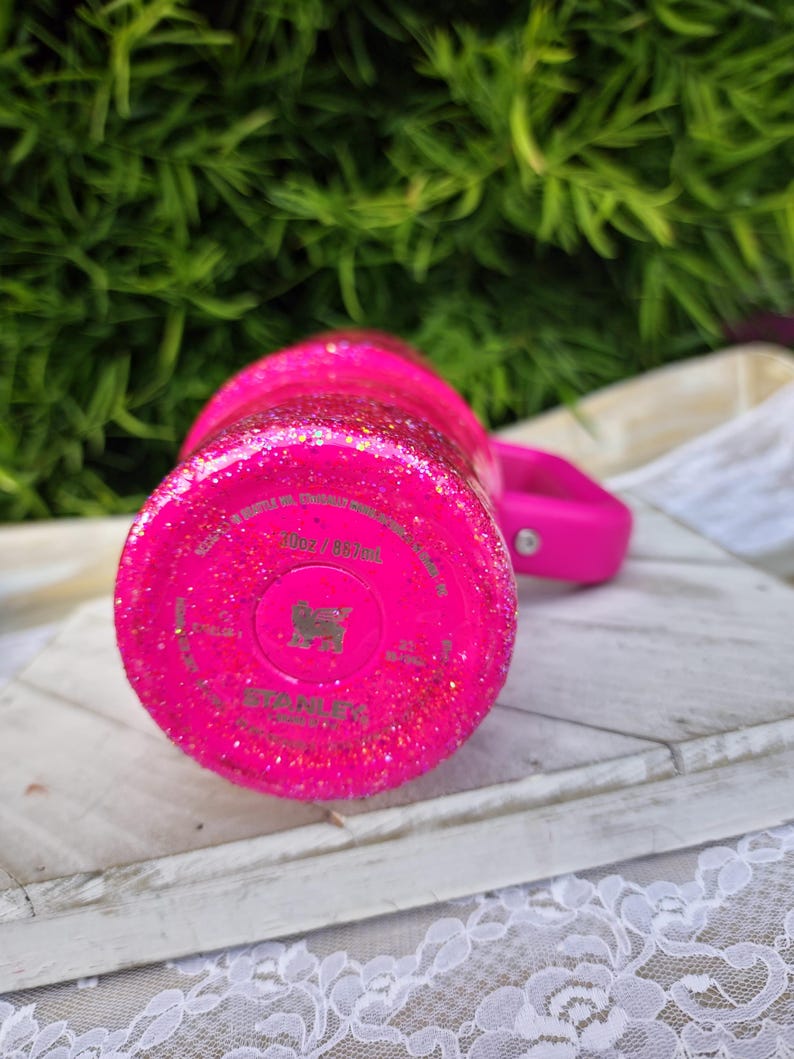 Pode incluir: Um copo rosa brilhante coberto de glitter com uma al&ccedil;a a condizer. A parte inferior do copo tem o nome e o log&oacute;tipo da marca. O copo est&aacute; sobre uma superf&iacute;cie de madeira branca com uma borda de renda branca.