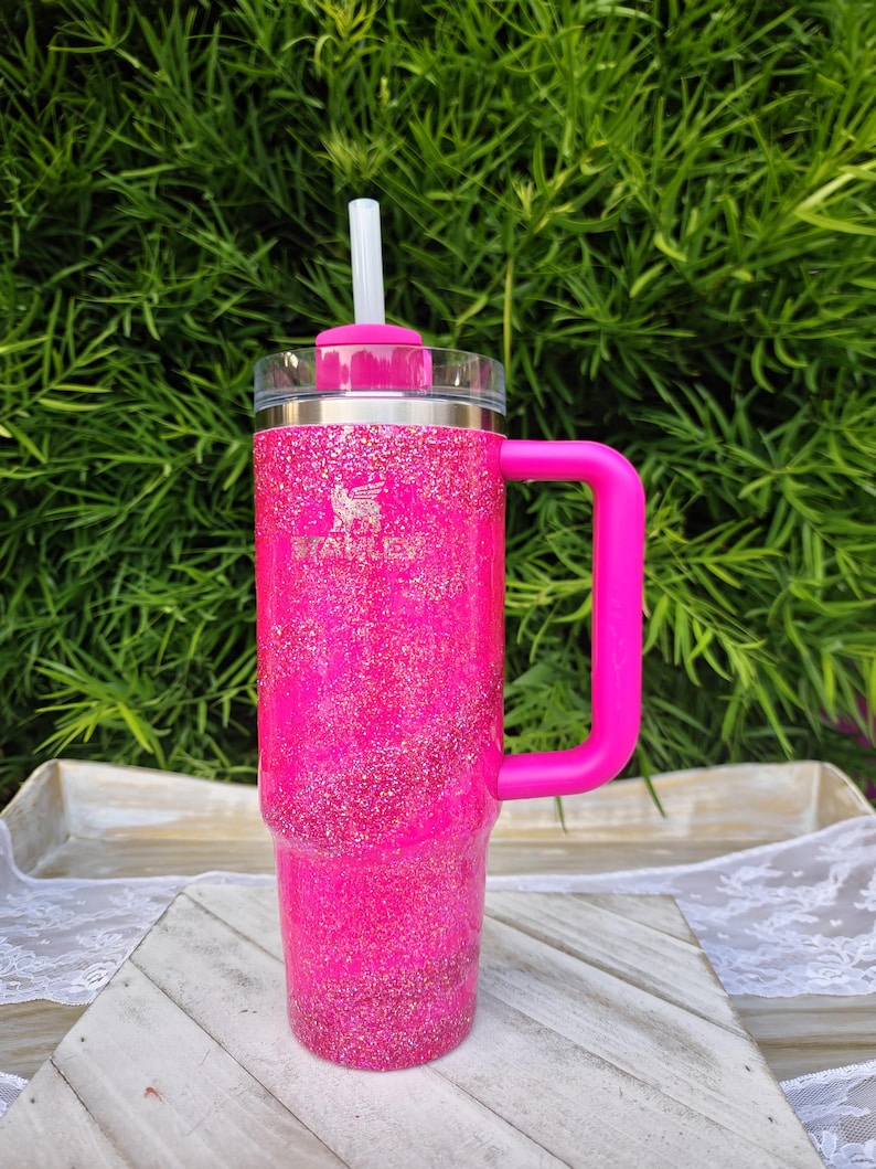 Pode incluir: Um copo rosa brilhante com glitter, com uma al&ccedil;a e canudo a condizer. O copo apresenta um log&oacute;tipo prateado e uma tampa transparente. A pega &eacute; rosa brilhante e o canudo &eacute; branco. Concebido para bebidas frias ou quentes.