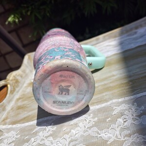 Puede incluir: Un vaso Stanley de 1,18 L con un acabado marmolado brillante en tonos rosa, azul y blanco. La base muestra el logotipo de Stanley, un icono de oso alado y el texto 40oz/1.18L. Un asa de color verde claro est&aacute; unida al lateral del vaso.