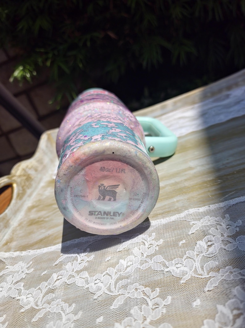 Puede incluir: Un vaso Stanley de 1,18 L con un acabado marmolado brillante en tonos rosa, azul y blanco. La base muestra el logotipo de Stanley, un icono de oso alado y el texto 40oz/1.18L. Un asa de color verde claro est&aacute; unida al lateral del vaso.