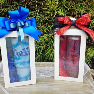 Puede incluir: Dos cajas de regalo blancas con ventanas transparentes, cada una con un vaso brillante. La caja de la izquierda presenta un vaso azul con el nombre Debbie y un lazo azul grande. La caja de la derecha contiene un vaso rojo con el nombre Amy Dean y un lazo rojo.