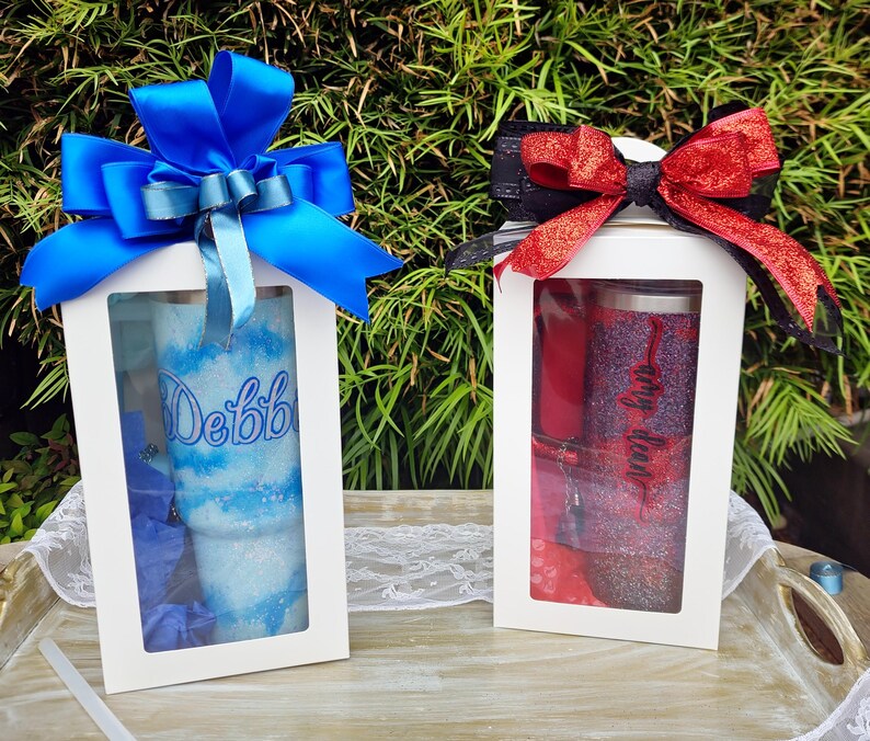 Puede incluir: Dos cajas de regalo blancas con ventanas transparentes, cada una con un vaso brillante. La caja de la izquierda presenta un vaso azul con el nombre Debbie y un lazo azul grande. La caja de la derecha contiene un vaso rojo con el nombre Amy Dean y un lazo rojo.