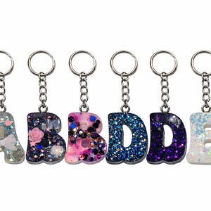 May include: Six glitter resin letter keychains spelling out the letters A, B, B, D, D, and E. Each letter is a different color and has a silver chain and keyring. The letters are filled with glitter and decorative elements.