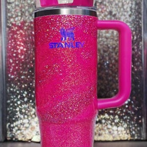 Pode incluir: Um copo Stanley rosa choque com glitter, com uma al&ccedil;a e tampa a condizer. O copo tem uma borda prateada e um canudo branco. O logotipo da Stanley &eacute; vis&iacute;vel em azul. O fundo &eacute; uma superf&iacute;cie brilhante.