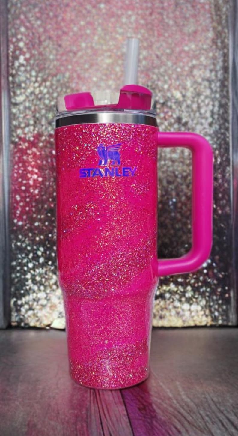 Pode incluir: Um copo Stanley rosa choque com glitter, com uma al&ccedil;a e tampa a condizer. O copo tem uma borda prateada e um canudo branco. O logotipo da Stanley &eacute; vis&iacute;vel em azul. O fundo &eacute; uma superf&iacute;cie brilhante.