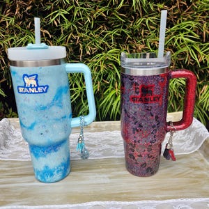 Puede incluir: Dos vasos de acero inoxidable con asas y pajitas. El vaso de la izquierda es azul claro con un acabado de purpurina marmolado y tapa blanca. El vaso de la derecha es rojo oscuro y negro con un estampado de leopardo y purpurina.