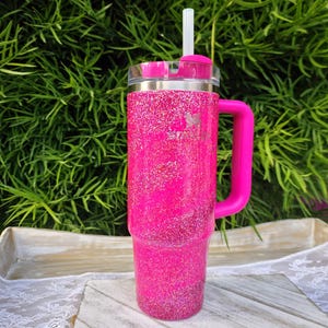 Pode incluir: Um copo rosa choque com glitter, com uma pega e uma tampa transparente. O copo tem uma faixa prateada e uma palhinha branca. A pega e a tampa tamb&eacute;m s&atilde;o rosa. Est&aacute; sobre uma superf&iacute;cie de madeira branca e cinzenta.