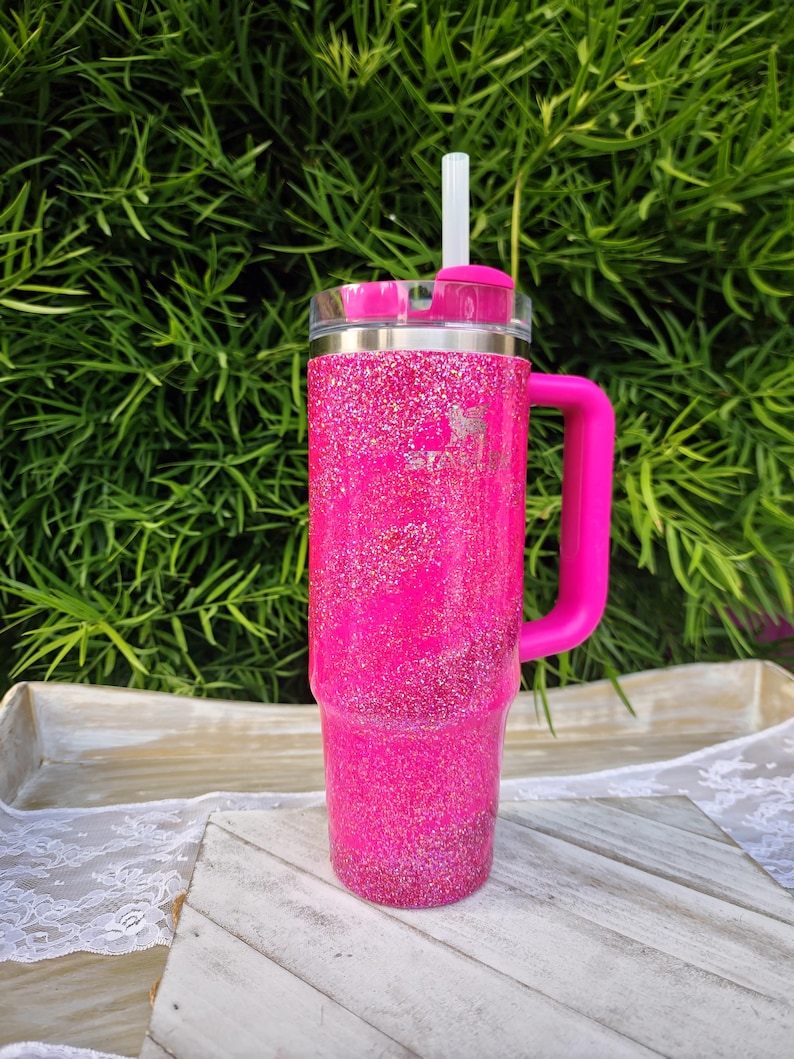 Pode incluir: Um copo rosa choque com glitter, com uma pega e uma tampa transparente. O copo tem uma faixa prateada e uma palhinha branca. A pega e a tampa tamb&eacute;m s&atilde;o rosa. Est&aacute; sobre uma superf&iacute;cie de madeira branca e cinzenta.