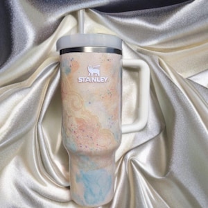 Puede incluir: Un vaso de la marca Stanley con asa y pajita. El vaso presenta una combinaci&oacute;n de colores pastel con una mezcla de tonos melocot&oacute;n, azul y amarillo. El logotipo de Stanley es visible en la parte delantera.
