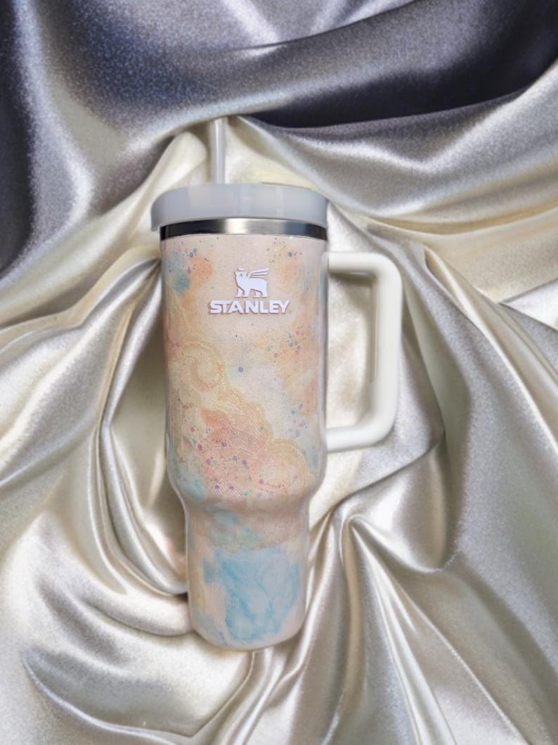 Puede incluir: Un vaso de la marca Stanley con asa y pajita. El vaso presenta una combinaci&oacute;n de colores pastel con una mezcla de tonos melocot&oacute;n, azul y amarillo. El logotipo de Stanley es visible en la parte delantera.