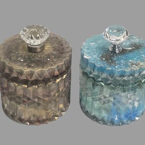 May include: Four decorative jars with faceted sides and crystal-like knobs. The jars are in shades of lavender, brown, blue, and dark blue, each with a glittery or textured appearance. The jars are cylindrical with a lid.