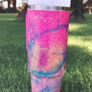 Puede incluir: Un vaso de acero inoxidable con un acabado de purpurina vibrante en rosa, azul y morado con un patr&oacute;n de remolinos. El vaso incluye una tapa blanca con un cierre deslizante de color rosa brillante y un asa rosa a juego en el lateral.