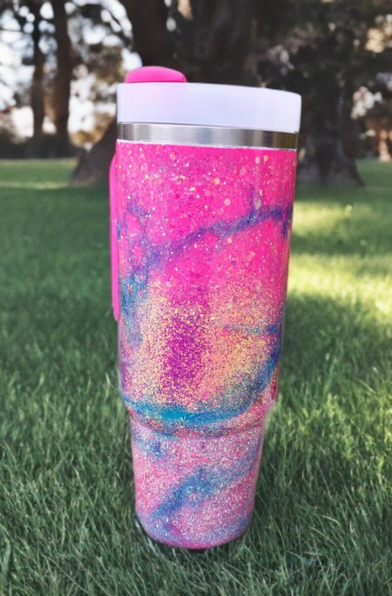 Puede incluir: Un vaso de acero inoxidable con un acabado de purpurina vibrante en rosa, azul y morado con un patr&oacute;n de remolinos. El vaso incluye una tapa blanca con un cierre deslizante de color rosa brillante y un asa rosa a juego en el lateral.