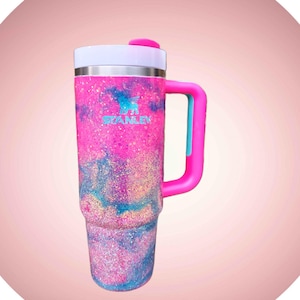 Puede incluir: Un vaso alto de acero inoxidable con asa, que presenta un acabado brillante con remolinos en rosa, azul y morado. El logotipo de Stanley est&aacute; impreso en azul claro en la parte frontal. Tiene una tapa blanca con una pesta&ntilde;a y asa de color rosa.