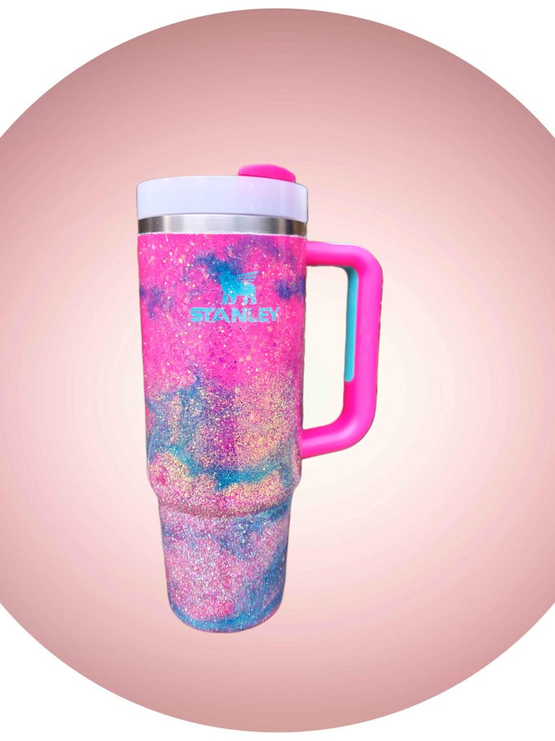 Puede incluir: Un vaso alto de acero inoxidable con asa, que presenta un acabado brillante con remolinos en rosa, azul y morado. El logotipo de Stanley est&aacute; impreso en azul claro en la parte frontal. Tiene una tapa blanca con una pesta&ntilde;a y asa de color rosa.
