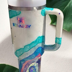 May include: A white Stanley tumbler with a handle and a white lid. The body features a glittery, multi-layered geode design in shades of teal, blue, and pink, with a holographic Stanley logo printed on the upper portion of the cup.