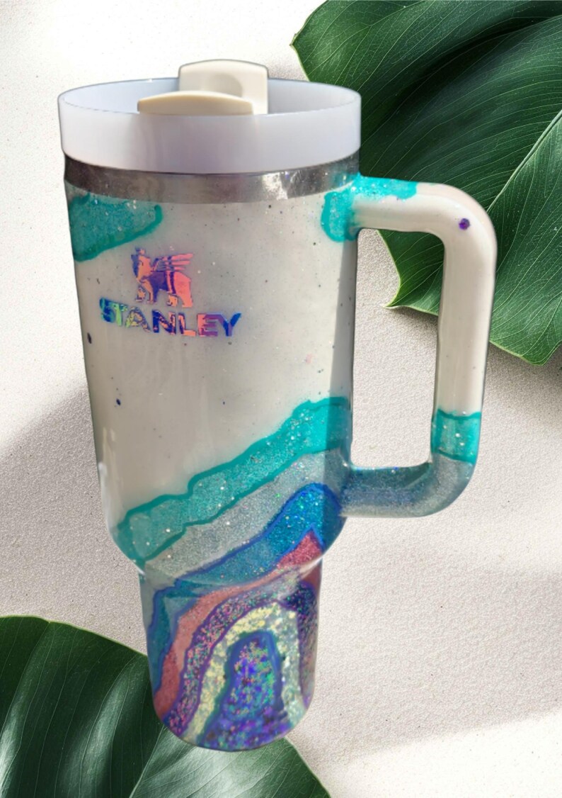 May include: A white Stanley tumbler with a handle and a white lid. The body features a glittery, multi-layered geode design in shades of teal, blue, and pink, with a holographic Stanley logo printed on the upper portion of the cup.