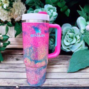 Puede incluir: Un vaso Stanley con un acabado brillante en tonos rosa, azul y dorado. El recipiente t&eacute;rmico incluye un asa lateral rosa resistente, una tapa blanca con un detalle rosa y el logotipo de Stanley impreso en color azul claro en la parte frontal.