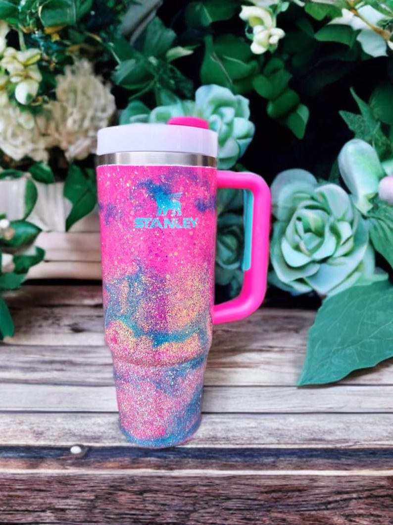 Puede incluir: Un vaso Stanley con un acabado brillante en tonos rosa, azul y dorado. El recipiente t&eacute;rmico incluye un asa lateral rosa resistente, una tapa blanca con un detalle rosa y el logotipo de Stanley impreso en color azul claro en la parte frontal.