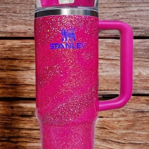 Pode incluir: Um copo Stanley rosa brilhante com uma al&ccedil;a e um canudo. O copo tem uma tampa e o logotipo Stanley em azul. A al&ccedil;a e a tampa s&atilde;o de cor rosa s&oacute;lida. O copo est&aacute; sobre um fundo de madeira.