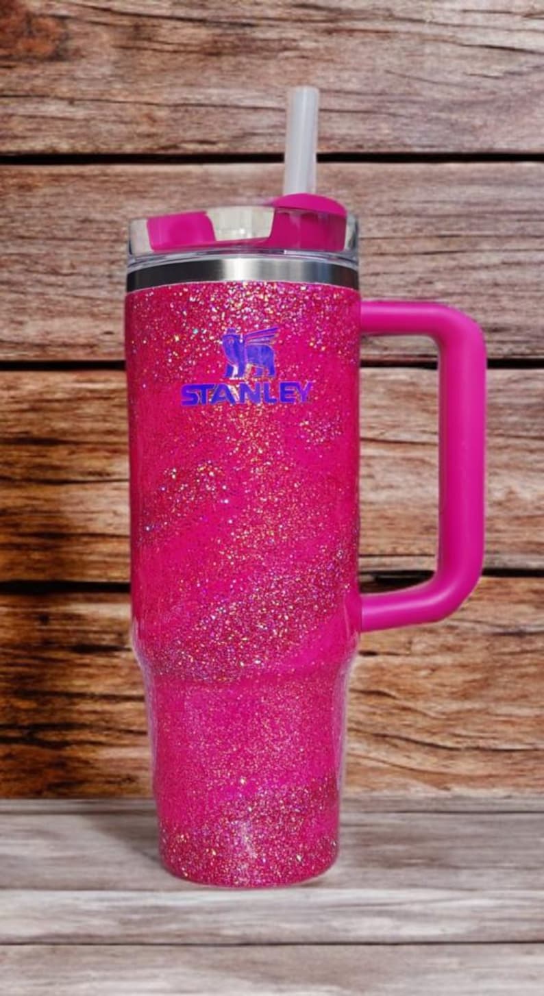 Pode incluir: Um copo Stanley rosa brilhante com uma al&ccedil;a e um canudo. O copo tem uma tampa e o logotipo Stanley em azul. A al&ccedil;a e a tampa s&atilde;o de cor rosa s&oacute;lida. O copo est&aacute; sobre um fundo de madeira.