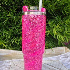 Pode incluir: Um copo rosa vibrante com glitter, tampa transparente e canudo rosa a condizer. O copo de a&ccedil;o inoxid&aacute;vel tem uma borda prateada. O fundo &eacute; uma folhagem verde desfocada.
