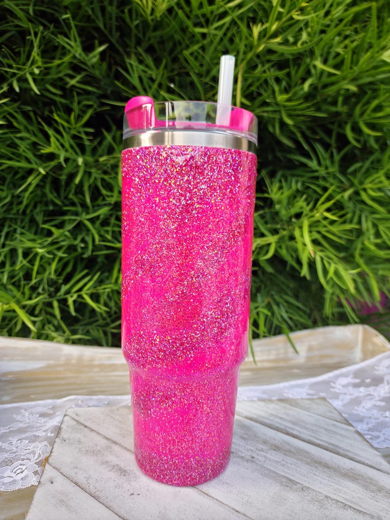 Pode incluir: Um copo rosa vibrante com glitter, tampa transparente e canudo rosa a condizer. O copo de a&ccedil;o inoxid&aacute;vel tem uma borda prateada. O fundo &eacute; uma folhagem verde desfocada.