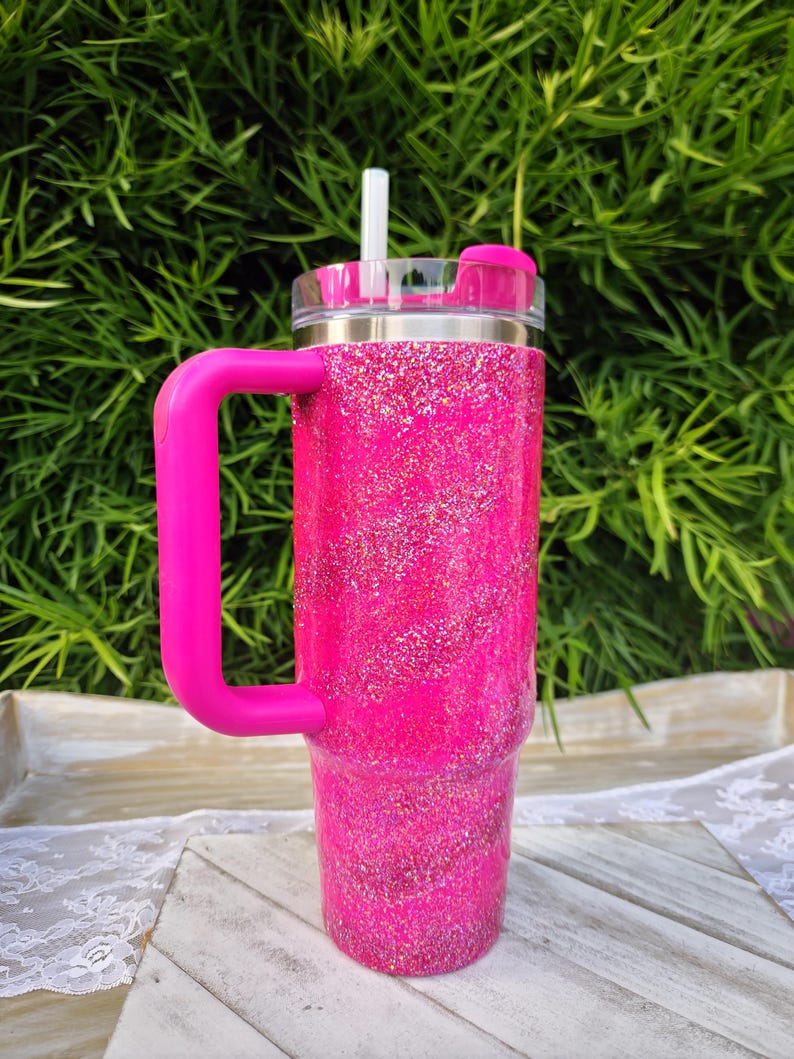 Pode incluir: Um copo rosa vibrante com glitter e uma al&ccedil;a e tampa a condizer. O copo tem uma tampa transparente com canudo e um controle deslizante rosa. A al&ccedil;a &eacute; de cor rosa brilhante. O copo est&aacute; em uma bandeja de madeira branca.