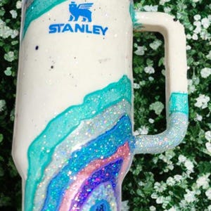May include: A white Stanley tumbler featuring a blue logo and a multi-colored geode-inspired design. The surface is decorated with swirling layers of teal, light blue, pink, and dark blue glitter, creating a textured, sparkling appearance.