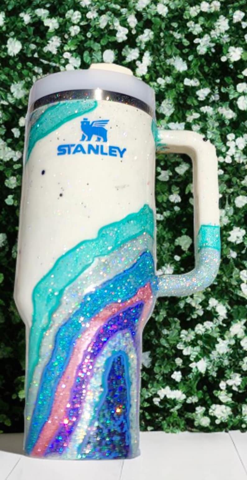 May include: A white Stanley tumbler featuring a blue logo and a multi-colored geode-inspired design. The surface is decorated with swirling layers of teal, light blue, pink, and dark blue glitter, creating a textured, sparkling appearance.