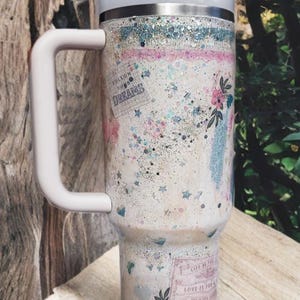 May include: A white tumbler with a handle, decorated with glitter, floral designs, and the words "Follow Your Dreams" and "Love is for Joy". The tumbler has a white lid and a reusable straw.