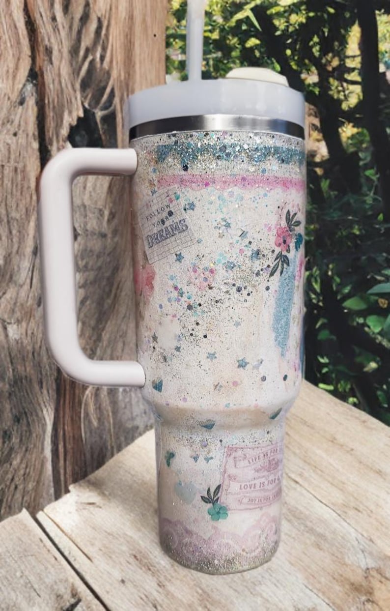 May include: A white tumbler with a handle, decorated with glitter, floral designs, and the words "Follow Your Dreams" and "Love is for Joy". The tumbler has a white lid and a reusable straw.