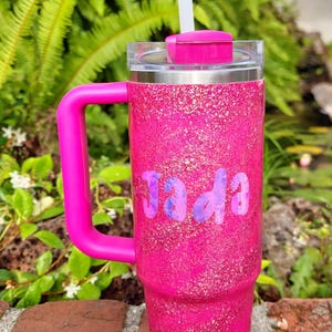 Puede incluir: Un vaso t&eacute;rmico de color rosa brillante con purpurina, asa rosa a juego, tapa transparente y pajita blanca. El nombre Jada est&aacute; impreso en el frente con una tipograf&iacute;a marmoleada en tonos morados y azules. El vaso tiene forma cil&iacute;ndrica.
