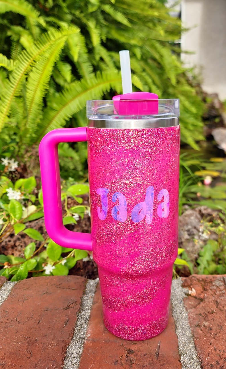 Puede incluir: Un vaso t&eacute;rmico de color rosa brillante con purpurina, asa rosa a juego, tapa transparente y pajita blanca. El nombre Jada est&aacute; impreso en el frente con una tipograf&iacute;a marmoleada en tonos morados y azules. El vaso tiene forma cil&iacute;ndrica.