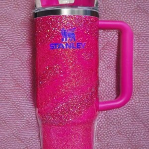 Pode incluir: Um copo Stanley rosa brilhante com uma al&ccedil;a e uma palhinha. O copo tem uma borda prateada e o logotipo Stanley em azul. A al&ccedil;a e a tampa s&atilde;o de cor rosa s&oacute;lida. O fundo &eacute; um tecido rosa texturizado.