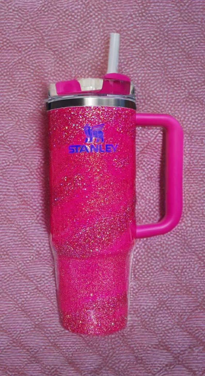 Pode incluir: Um copo Stanley rosa brilhante com uma al&ccedil;a e uma palhinha. O copo tem uma borda prateada e o logotipo Stanley em azul. A al&ccedil;a e a tampa s&atilde;o de cor rosa s&oacute;lida. O fundo &eacute; um tecido rosa texturizado.