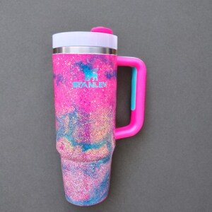 Puede incluir: Un vaso Stanley con un acabado de purpurina vibrante en rosa, azul y morado con un patr&oacute;n de m&aacute;rmol. El vaso incluye una tapa blanca con una pesta&ntilde;a rosa, un asa rosa brillante con un acento azul y el logotipo de Stanley impreso en azul.