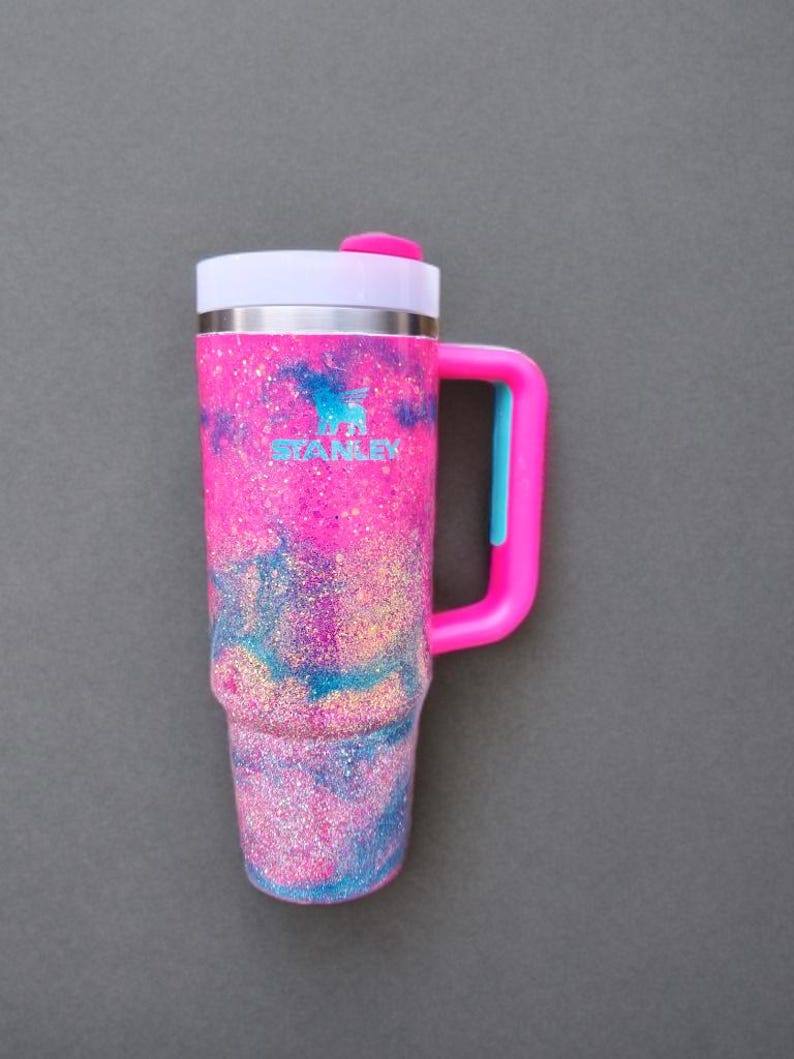 Puede incluir: Un vaso Stanley con un acabado de purpurina vibrante en rosa, azul y morado con un patr&oacute;n de m&aacute;rmol. El vaso incluye una tapa blanca con una pesta&ntilde;a rosa, un asa rosa brillante con un acento azul y el logotipo de Stanley impreso en azul.
