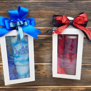 Puede incluir: Dos cajas de regalo blancas con ventanas transparentes, cada una con un vaso brillante. La caja de la izquierda presenta un vaso azul y blanco con el nombre Debbie y un gran lazo de sat&eacute;n azul. La caja de la derecha contiene un vaso rojo y negro con el nombre Amy y un lazo rojo brillante.