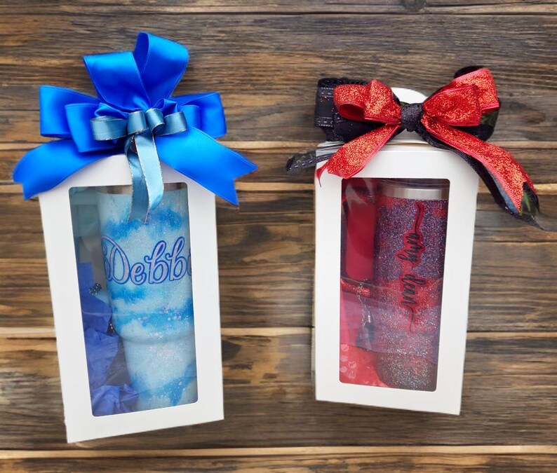 Puede incluir: Dos cajas de regalo blancas con ventanas transparentes, cada una con un vaso brillante. La caja de la izquierda presenta un vaso azul y blanco con el nombre Debbie y un gran lazo de sat&eacute;n azul. La caja de la derecha contiene un vaso rojo y negro con el nombre Amy y un lazo rojo brillante.
