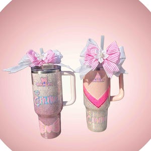 Puede incluir: Dos vasos de acero inoxidable con asa y pajita, con acabados brillantes en rosa y blanco. Cada vaso muestra el logotipo de Stanley, el nombre Emme en letra cursiva y un gran lazo decorativo con rayas rosas y detalles blancos en la tapa.