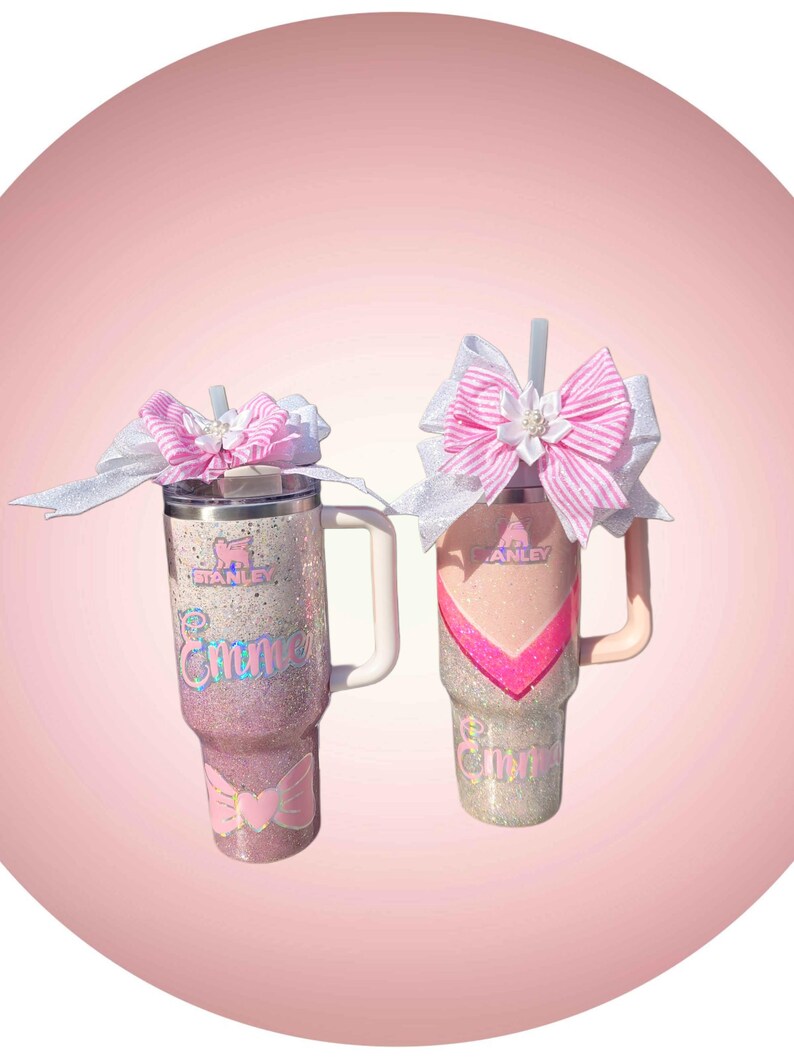 Puede incluir: Dos vasos de acero inoxidable con asa y pajita, con acabados brillantes en rosa y blanco. Cada vaso muestra el logotipo de Stanley, el nombre Emme en letra cursiva y un gran lazo decorativo con rayas rosas y detalles blancos en la tapa.